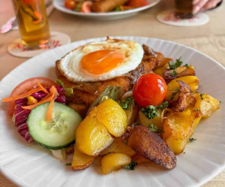 Gebratenes Schnitzel mit Spiegelei, Bratkartoffeln und kleinem Salat, serviert auf einem Teller im Brauhaus, mit zwei Gläsern Kölsch im Hintergrund